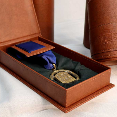 Бронзов медал със синя лента в отворена кафява кожена кутия. Отзад се вижда кожен тубус с релефен надпис