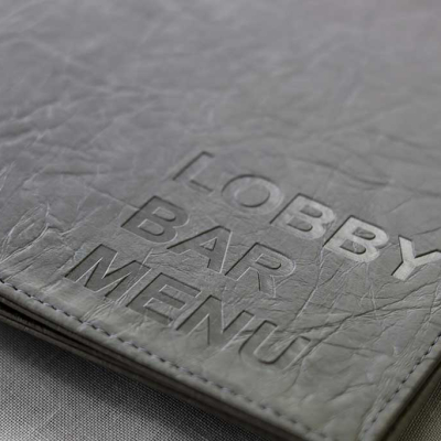 Релефен надпис lobby bar menu върху папка от еко кожа в сив цвят. По края има шев със светло сив конец.