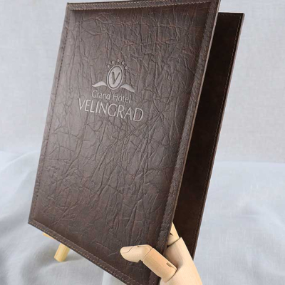 Кафява папка/меню с текстурирана кожа, върху която е релефно изобразено лого Grand Hotel VELINGRAD, поддържана от дървена ръка.